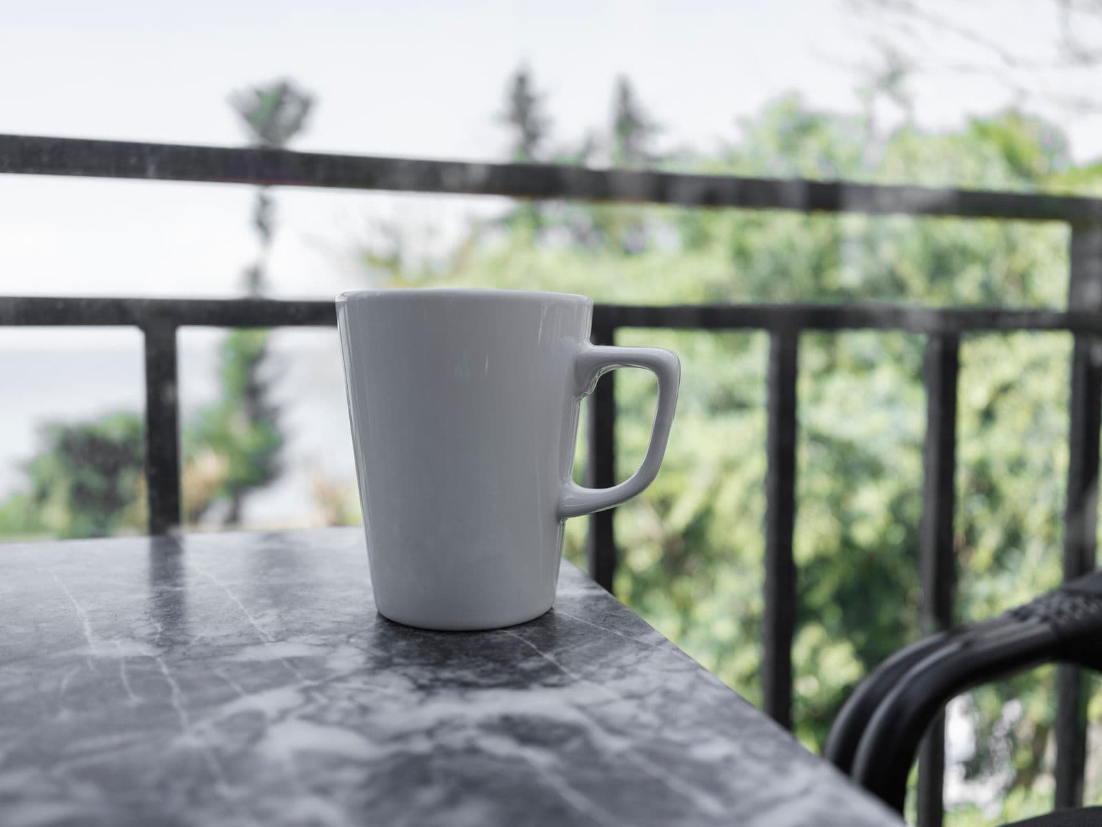 A white coffee mug on a marble table on a balcony, with green trees and the water beyond