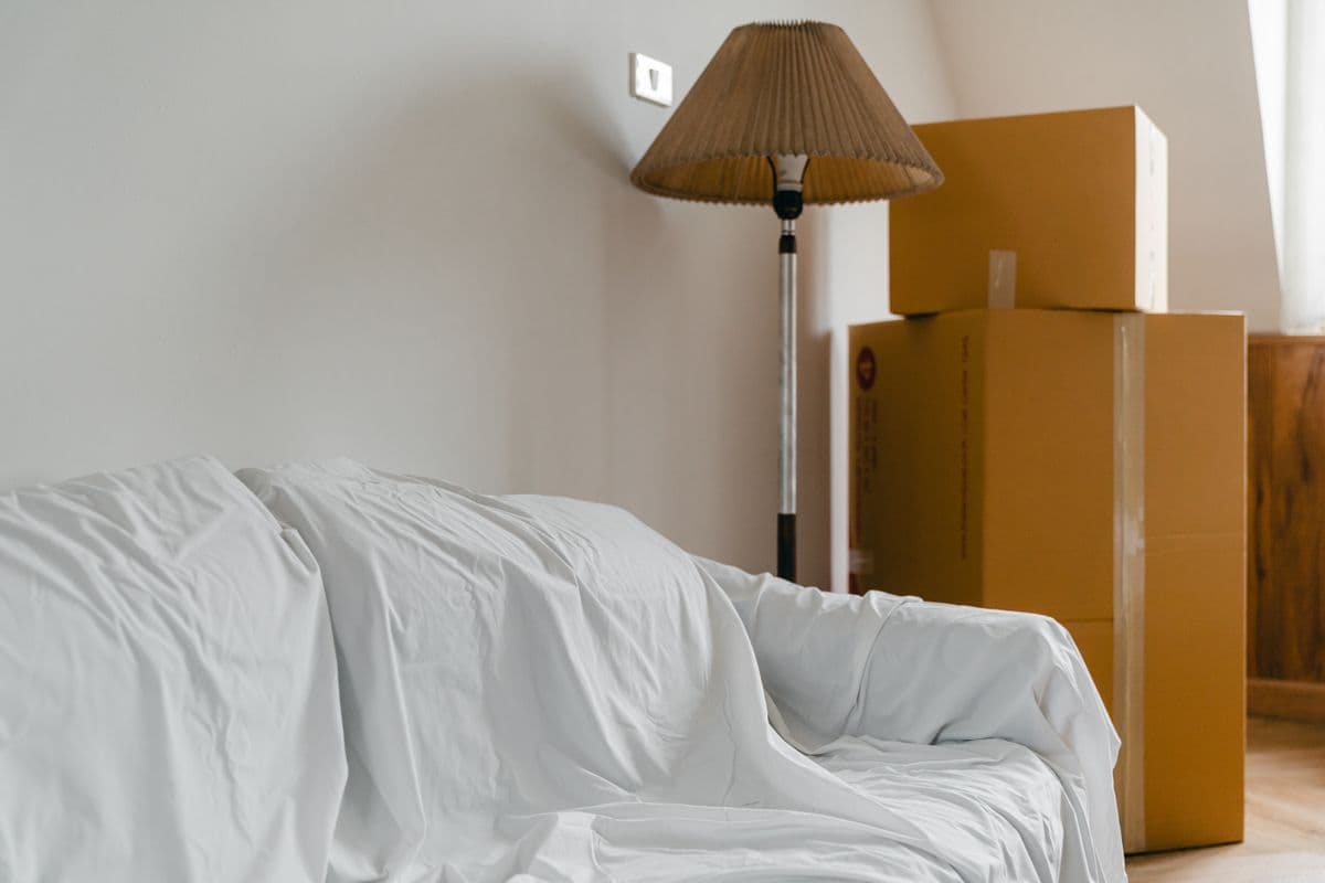 A sofa covered in a white sheet beside a floor lamp and stacked cardboard boxes in a bright room