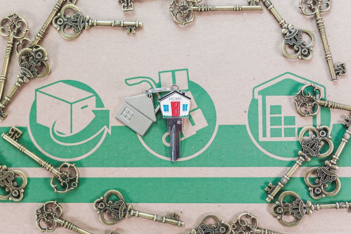 Antique skeleton keys arranged around a small house-shaped keychain on a cardboard surface printed with moving and home icons